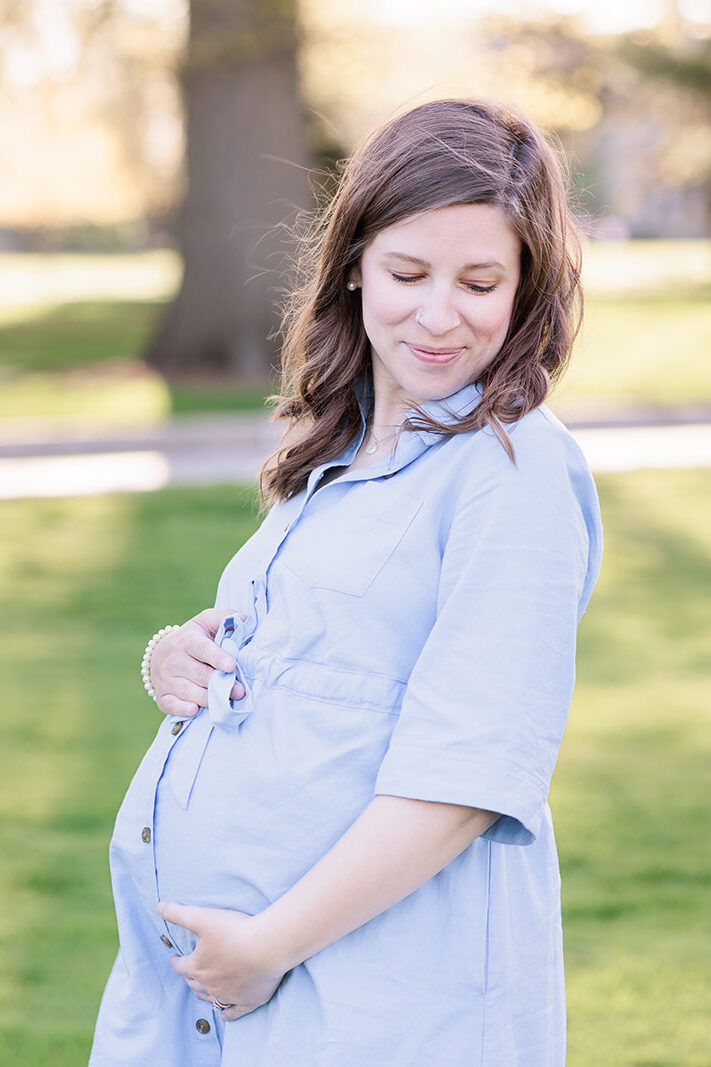 Pregnant woman holds her stomach as she looks away from the camera at Haverford arboretum posing for family photo session.