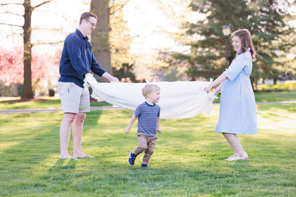 Candid photo of toddler running under blanket while father and mother hold white blanket 