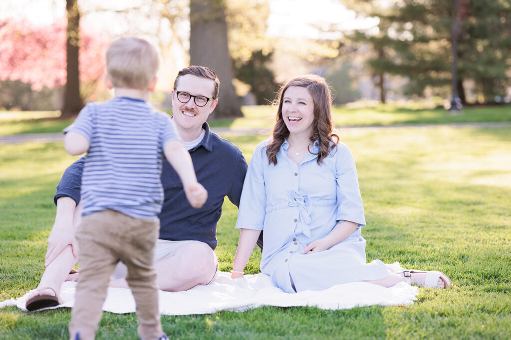 Toddler runs to parents seated on blanket at Haverford arboretum for family photo session on Main Line