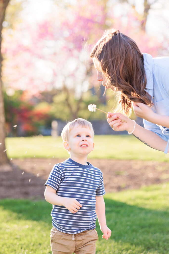 Mother blows dandelion as toddler looks on in awe at Main Line area arboretum 