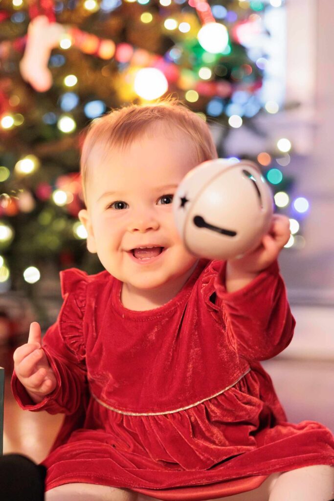 Baby holds up bell in front of Christmas tree during in-home baby photography session