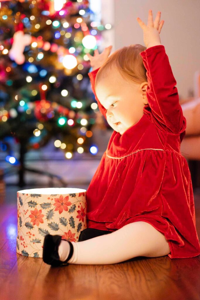 Baby raises her arms looking at glowing box in front of Christmas tree