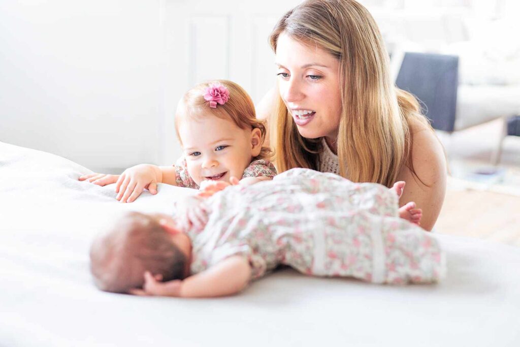 Young girl and mother look at newborn baby on bed, in-home newborn photo session Main Line, Philadelphia