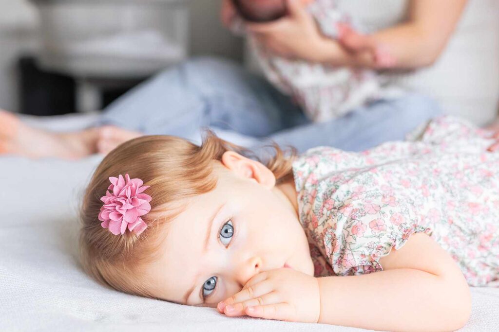 Toddler sucks her thumb and looks to camera during newborn photography session.