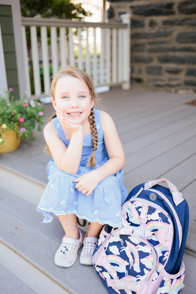 Girl poses on porch with backpack for first day of school photo.