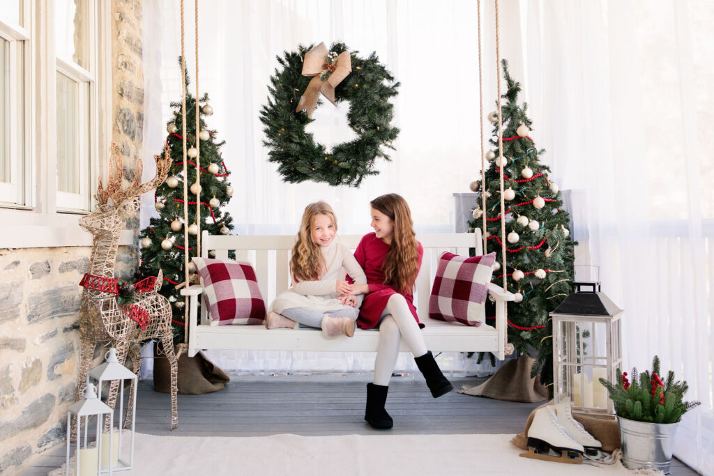 Two girls sit on porch swing surrounded by holiday decorations during main line holiday mini session